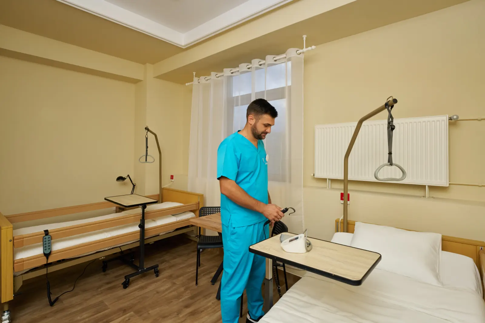 Asistent medical în scrubs turcoaz stând între două saltele medicale într-o cameră, AsertivO Otopeni