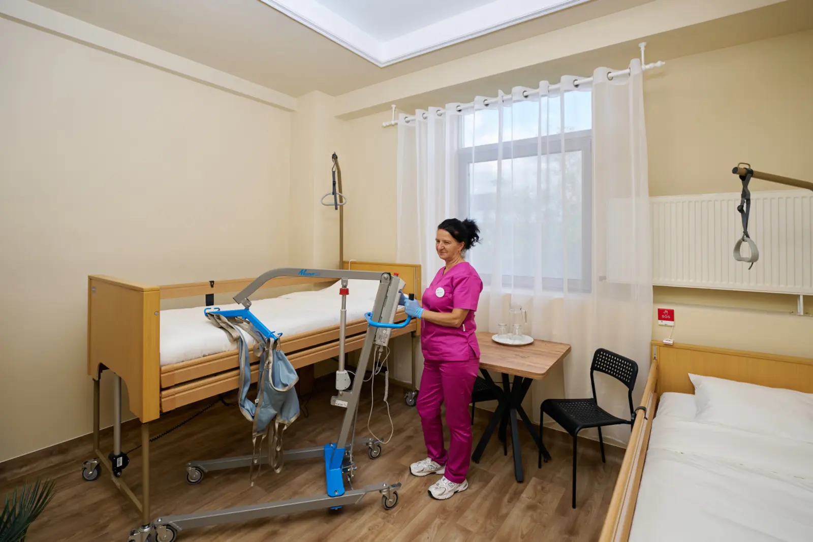 Asistentă medicală în scrub roz manevrează un lift electric de transfer rezident lângă un saltea medical, AsertivO Otopeni
