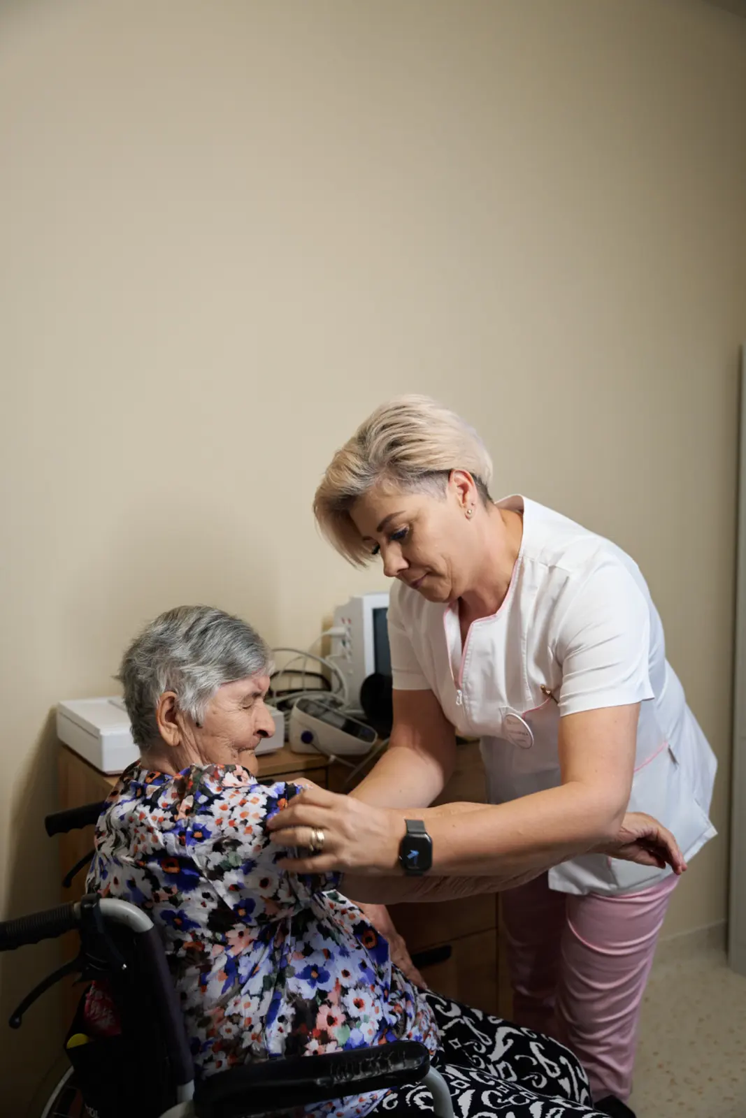 Asistentă medicală în uniformă albă ajută o senioră cu păr grizonant așezată în scaun rulant, AsertivO Otopeni