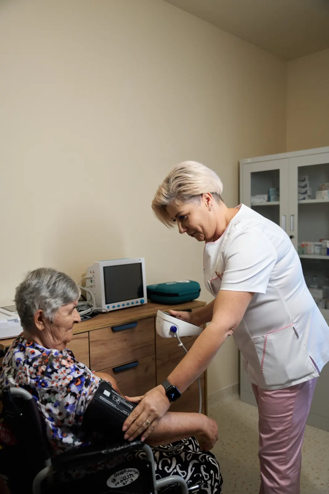 Asistentă medicală în uniformă albă cu roz măsoară tensiunea arterială unei doamne în vârstă așezată, AsertivO Otopeni