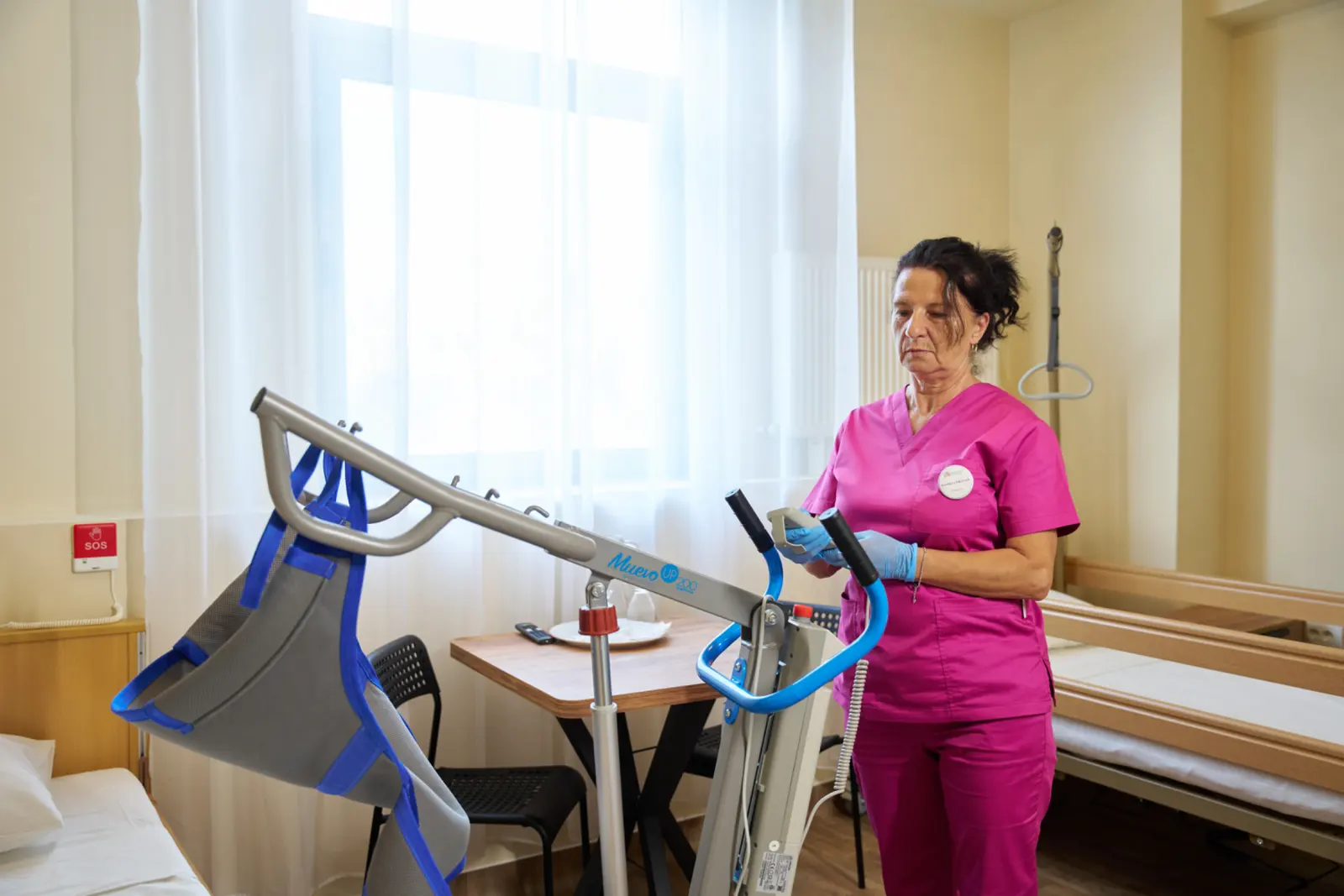 Asistentă medicală în uniformă roz operând un lift electric de transfer rezidenți Muevo UP într-o cameră, AsertivO Otopeni