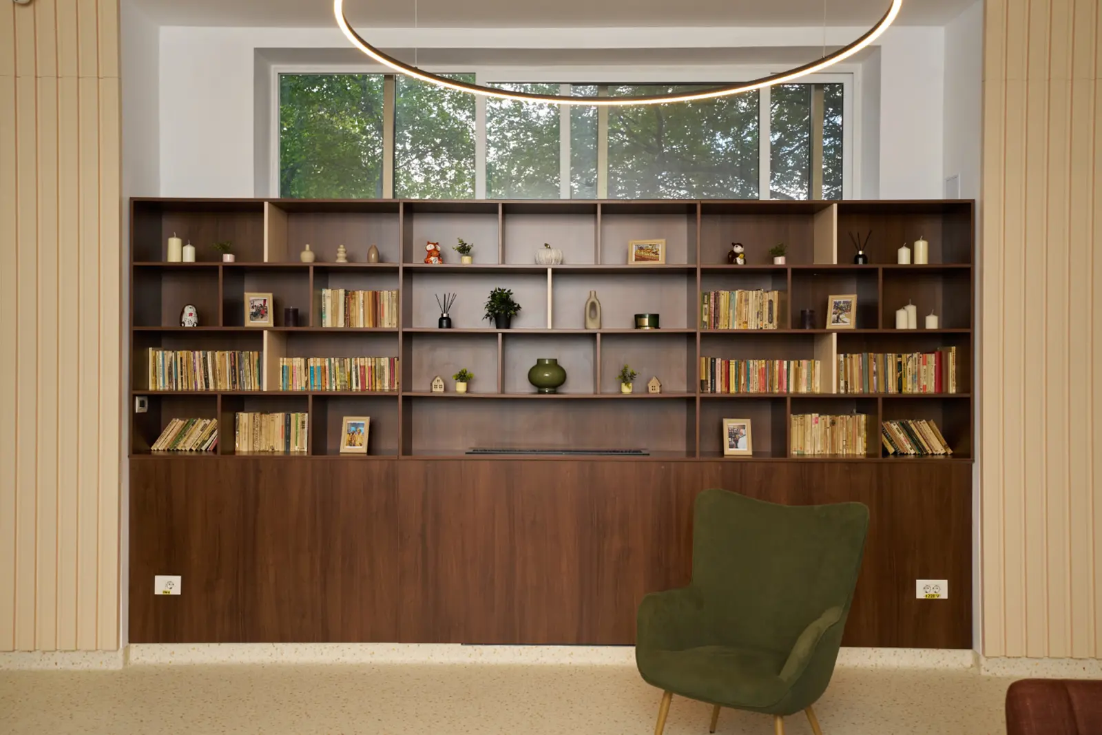 Bibliotecă mare din lemn închis cu cărți, decorațiuni și plante, fotoliu verde confortabil, AsertivO Otopeni