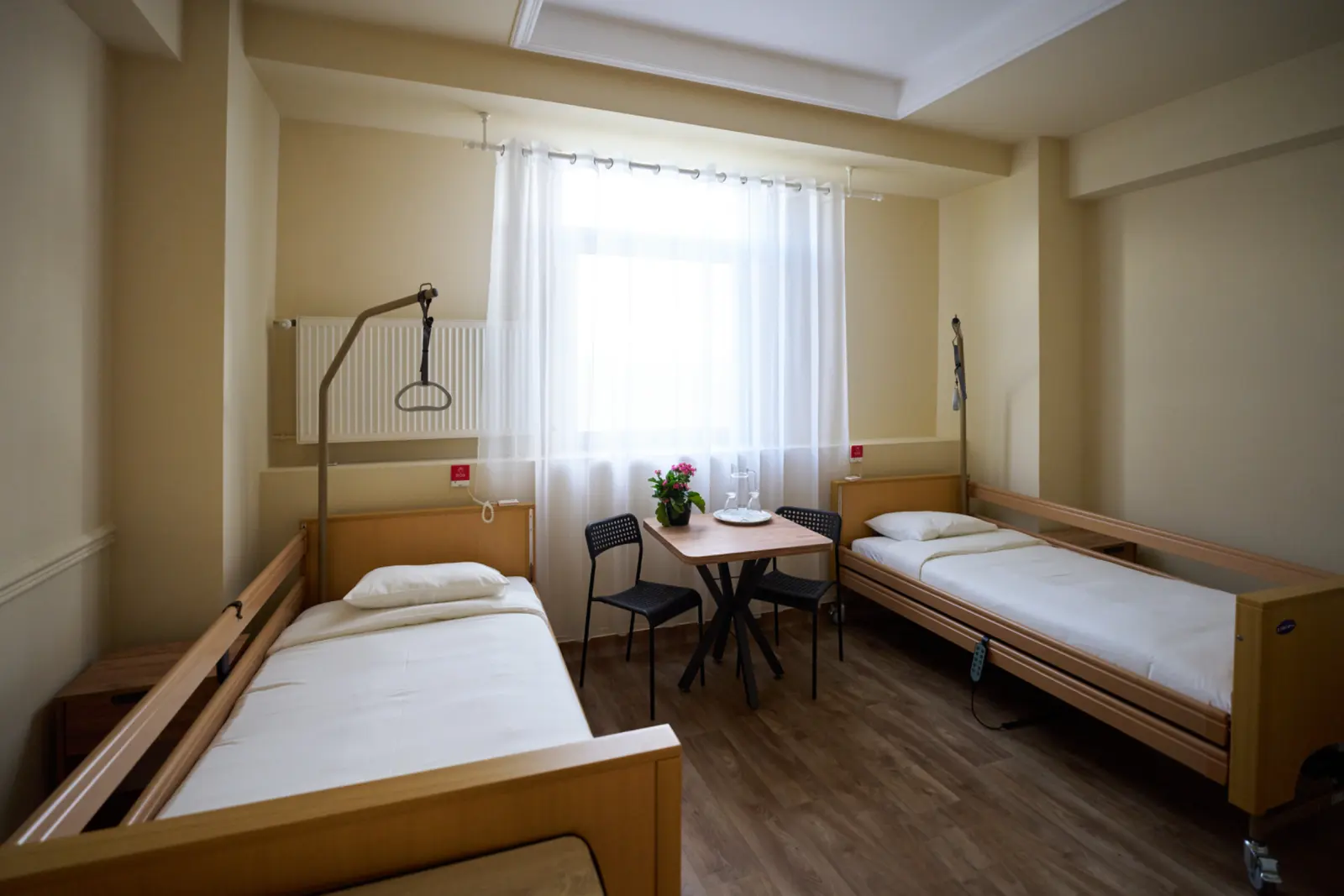 Cameră dublă dintr-un centru de îngrijire seniori cu două saltele medicale din lemn, lenjerie albă, AsertivO Otopeni
