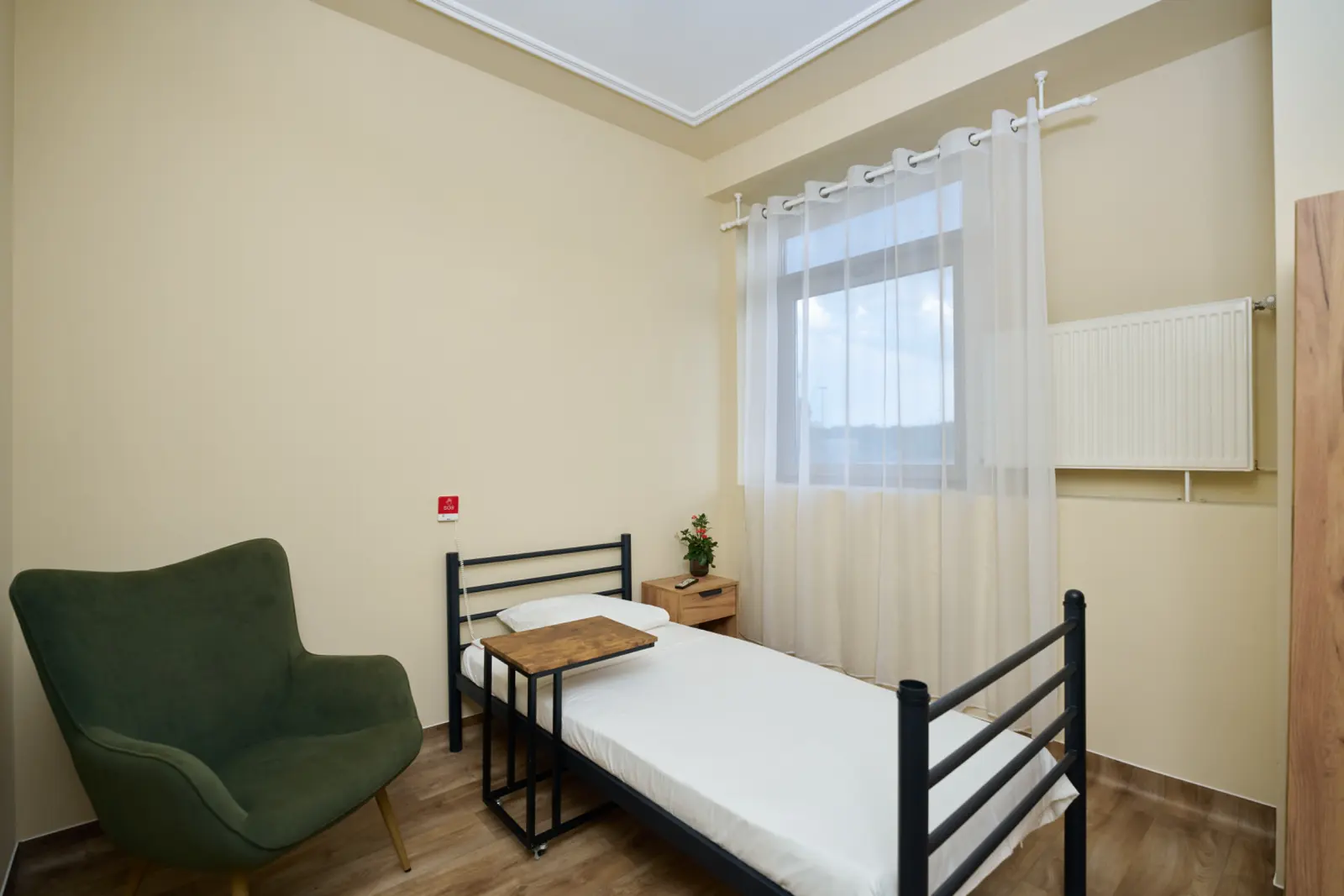 Cameră single pentru seniori cu saltea cu cadru metalic negru, fotoliu verde, noptieră din lemn, AsertivO Otopeni