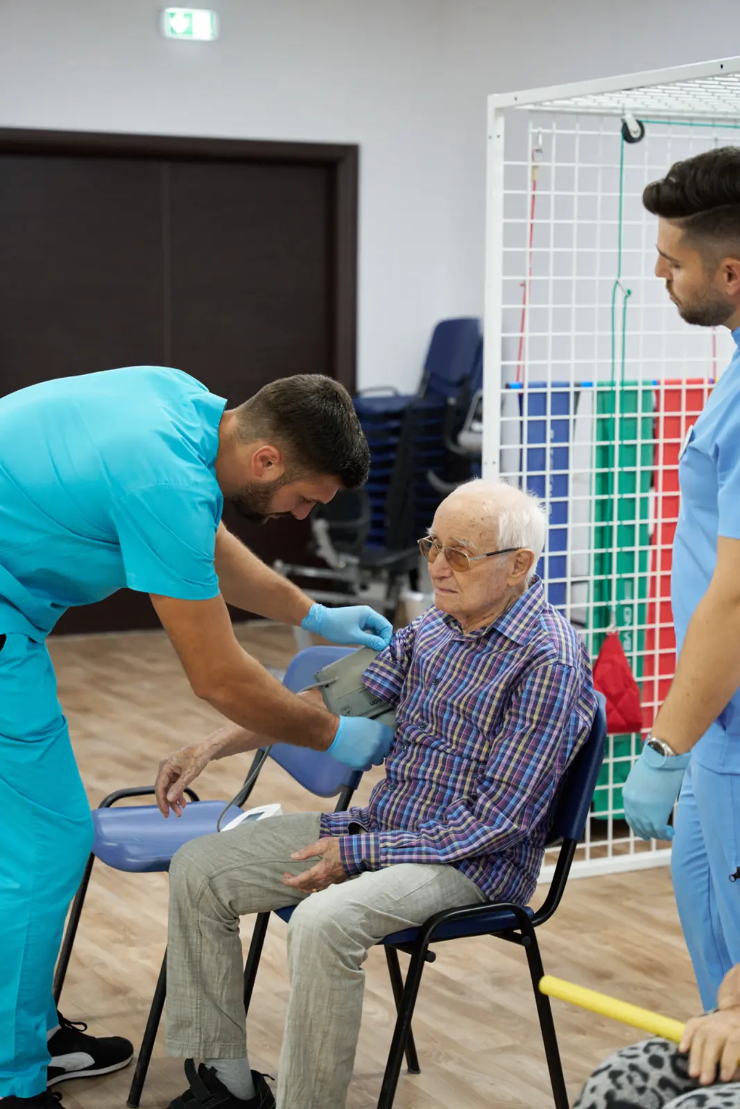 Doi asistenți medicali în uniformă bleu măsoară tensiunea arterială unui senior cu ochelari, AsertivO Otopeni