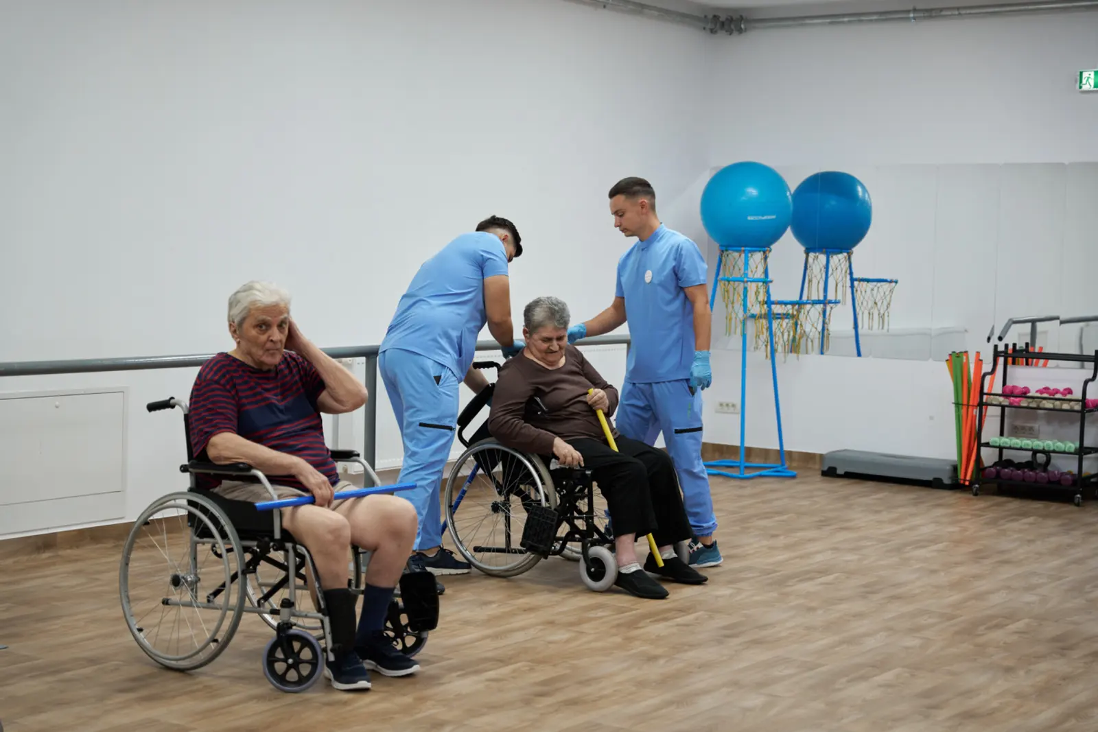 Doi kinetoterapeuți în uniforme albastre ajută doi rezidenți vârstnici în scaune, AsertivO Otopeni