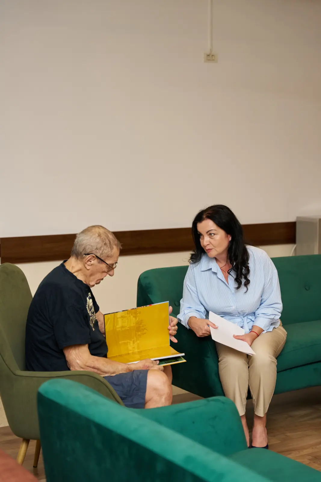 O femeie în cămașă albastră stă pe o canapea verde alături de un bărbat în vârstă care ține o carte, AsertivO Otopeni