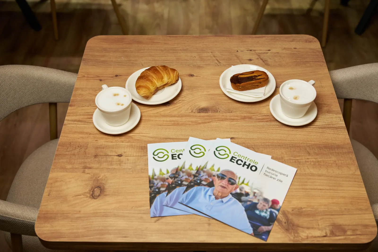 Masă de lemn cu două cafele cu lapte, un croissant, un ecler glazurat și broșuri promoționale Centrele ECHO, AsertivO Otopeni