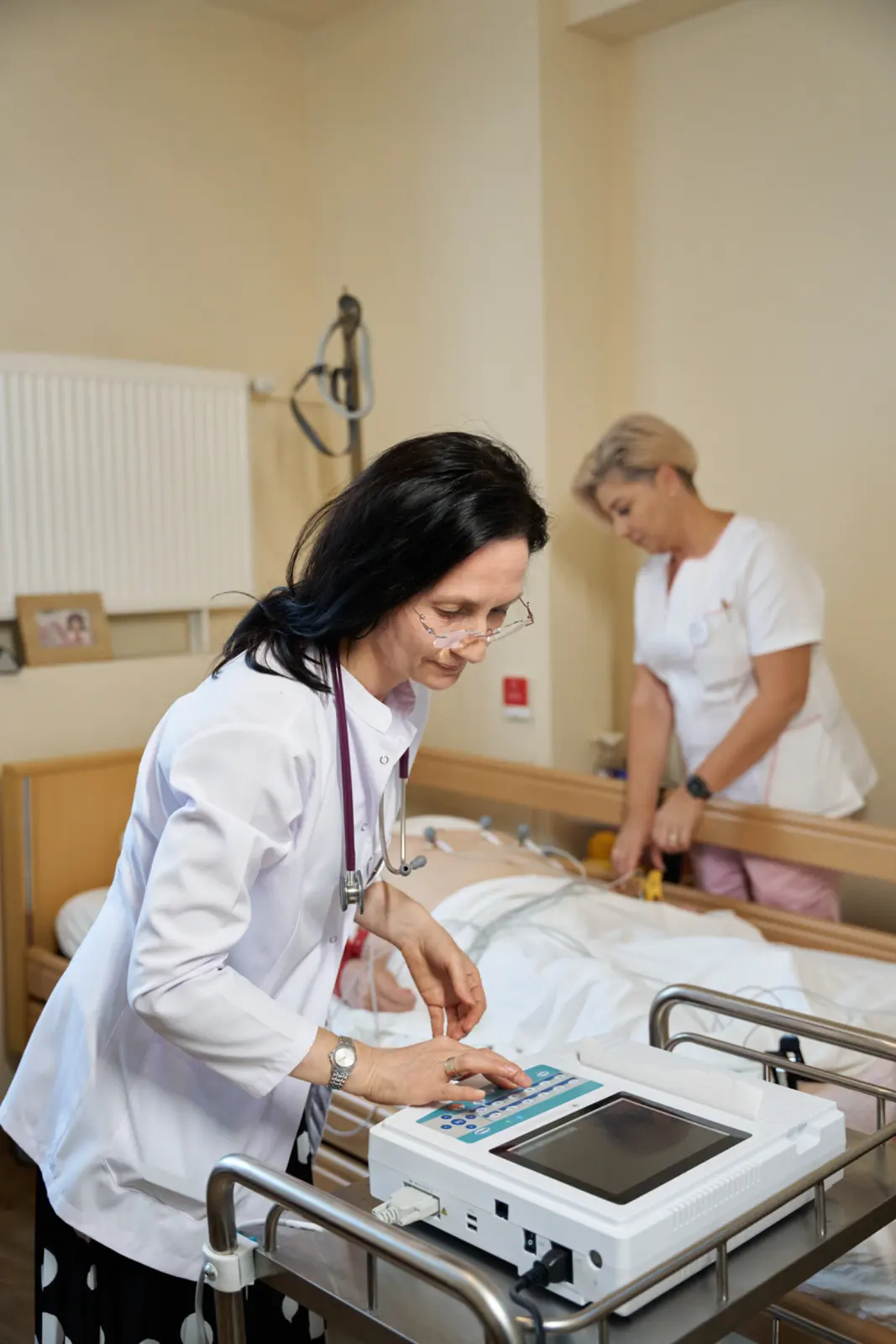 Medic în halat alb cu stetoscop operează un aparat ECG lângă salteaua unui rezident, AsertivO Otopeni
