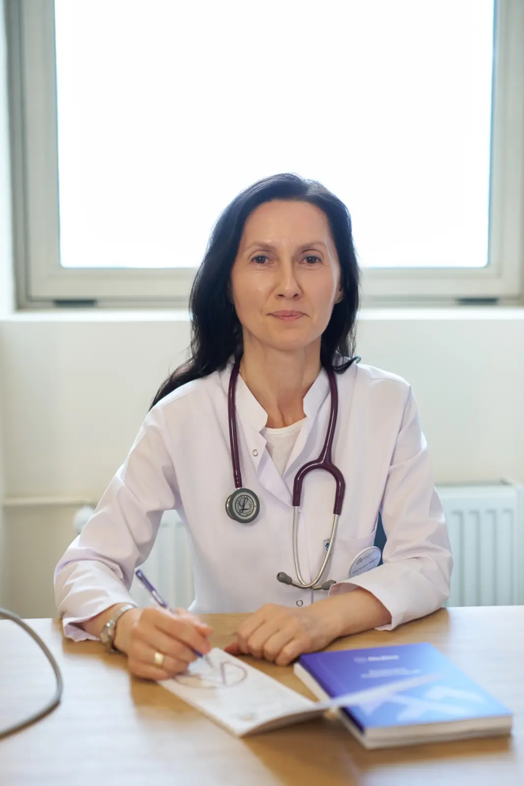 Medic specialist femeie în halat alb cu stetoscop, așezată la birou, consultând documente medicale, AsertivO Otopeni