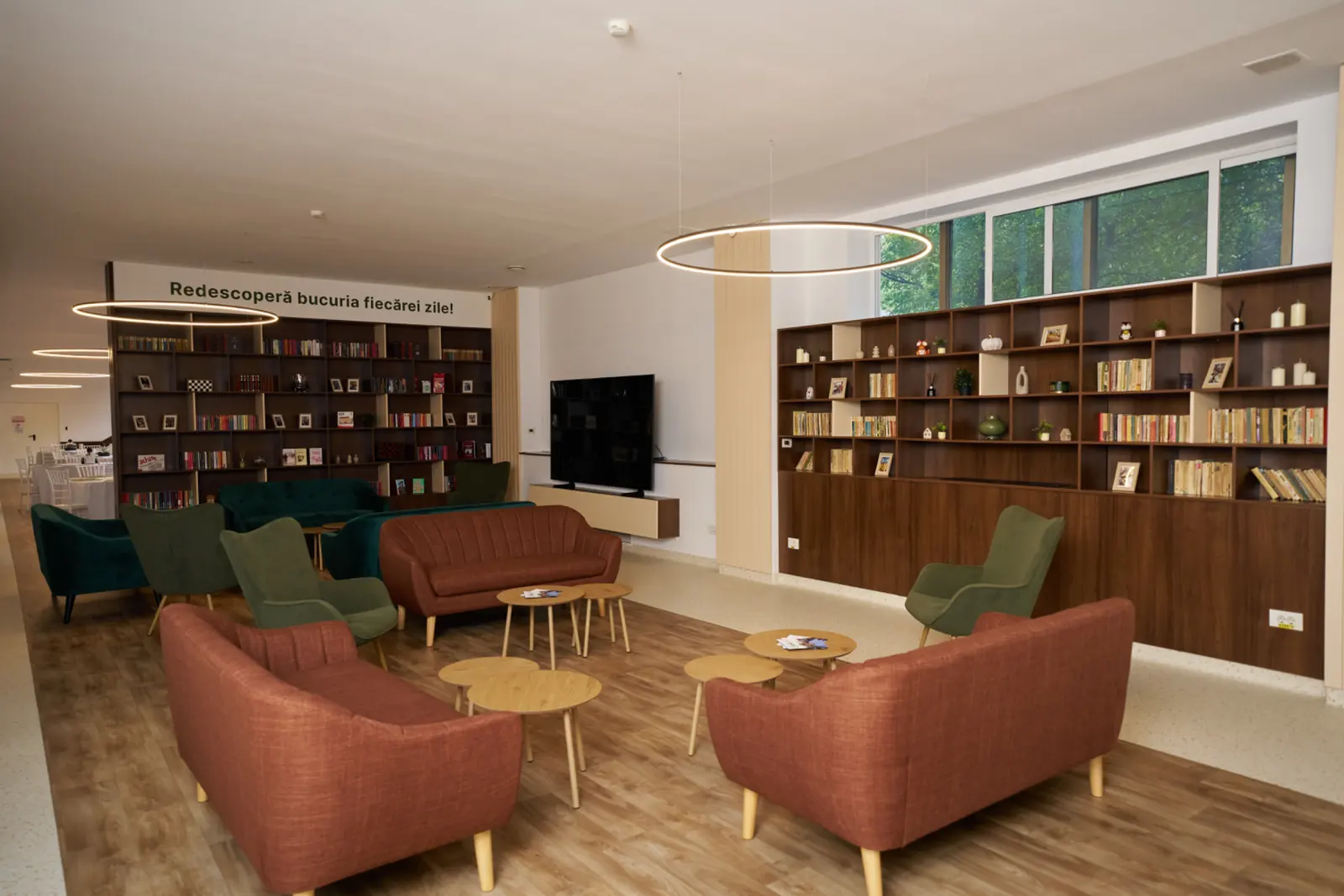 Salon elegant de relaxare cu biblioteci din lemn, fotolii roșii și verzi, mese rotunde din lemn, AsertivO Otopeni