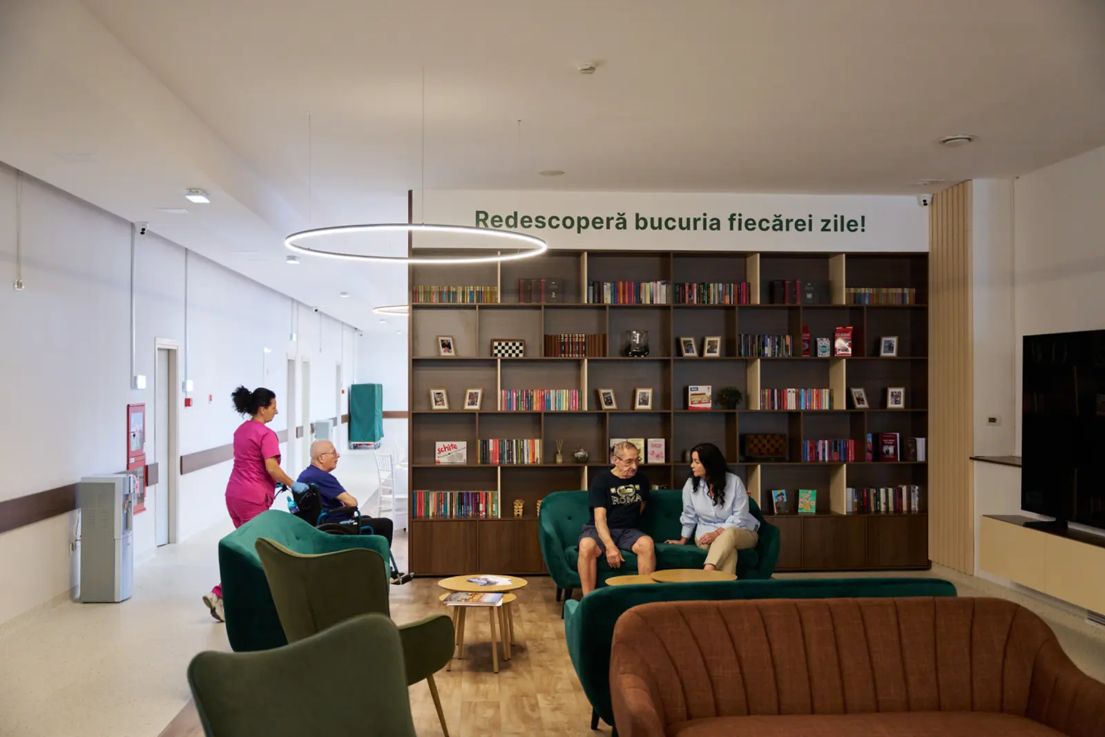 Salon de relaxare cu bibliotecă mare, fotolii verzi și maro, unde un senior conversează, AsertivO Otopeni