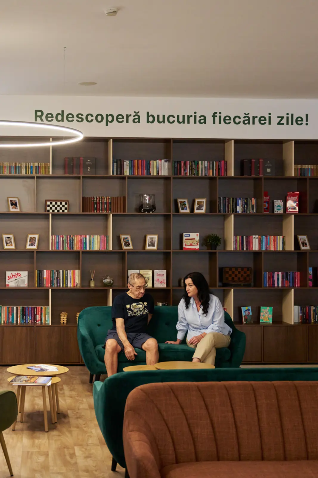 Un senior și o îngrijitoare stând de vorbă pe o canapea verde într-un centru de îngrijire seniori, AsertivO Otopeni