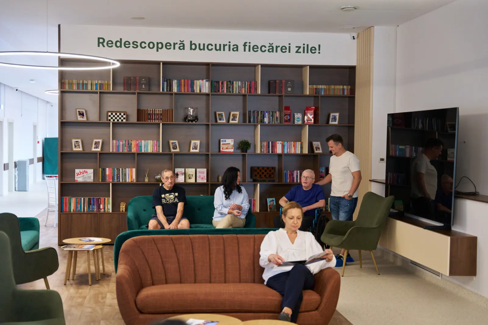 Seniori și personal în sala de relaxare a unui centru de îngrijire, cu bibliotecă mare în fundal, AsertivO Otopeni