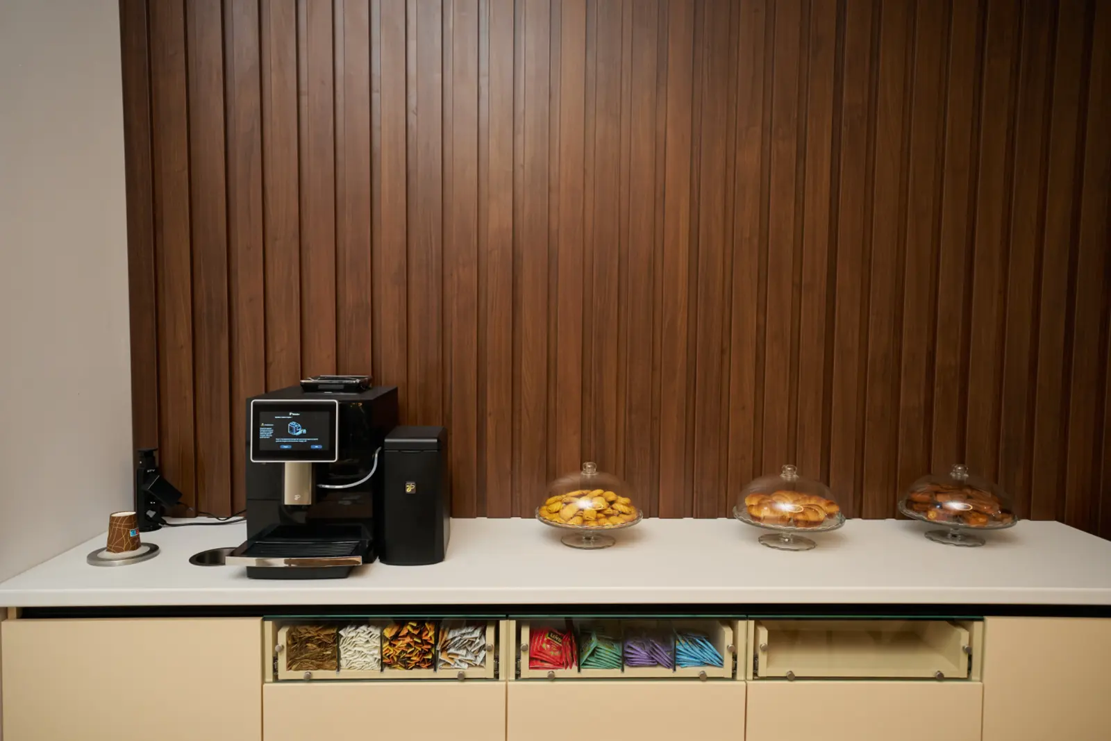 Tejghea albă cu espressor automat negru și trei platouri de sticlă cu produse de patiserie, AsertivO Otopeni