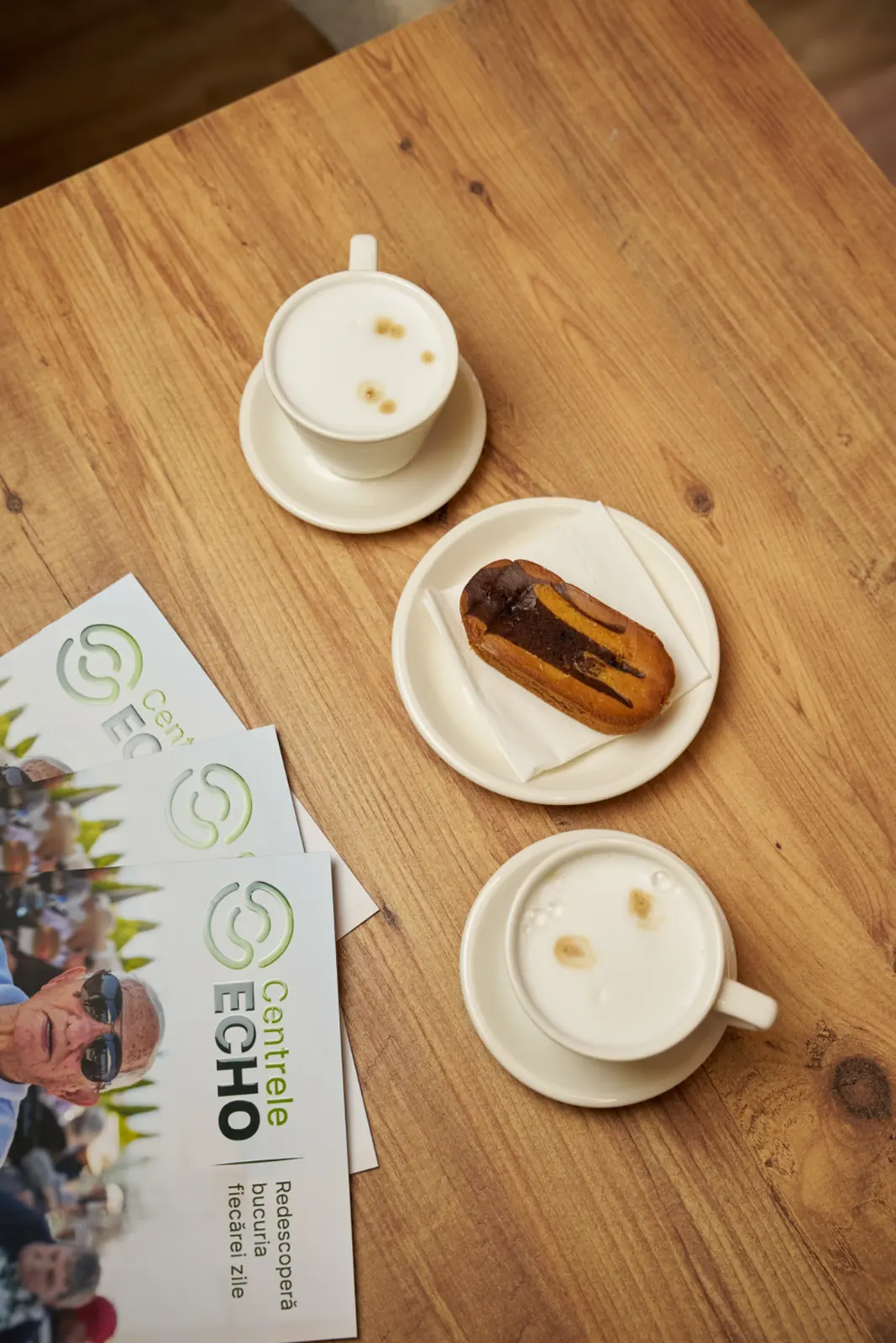 Vedere de sus a unei mese de lemn cu două căni de cappuccino pe farfurioare albe, un ecler, AsertivO Otopeni