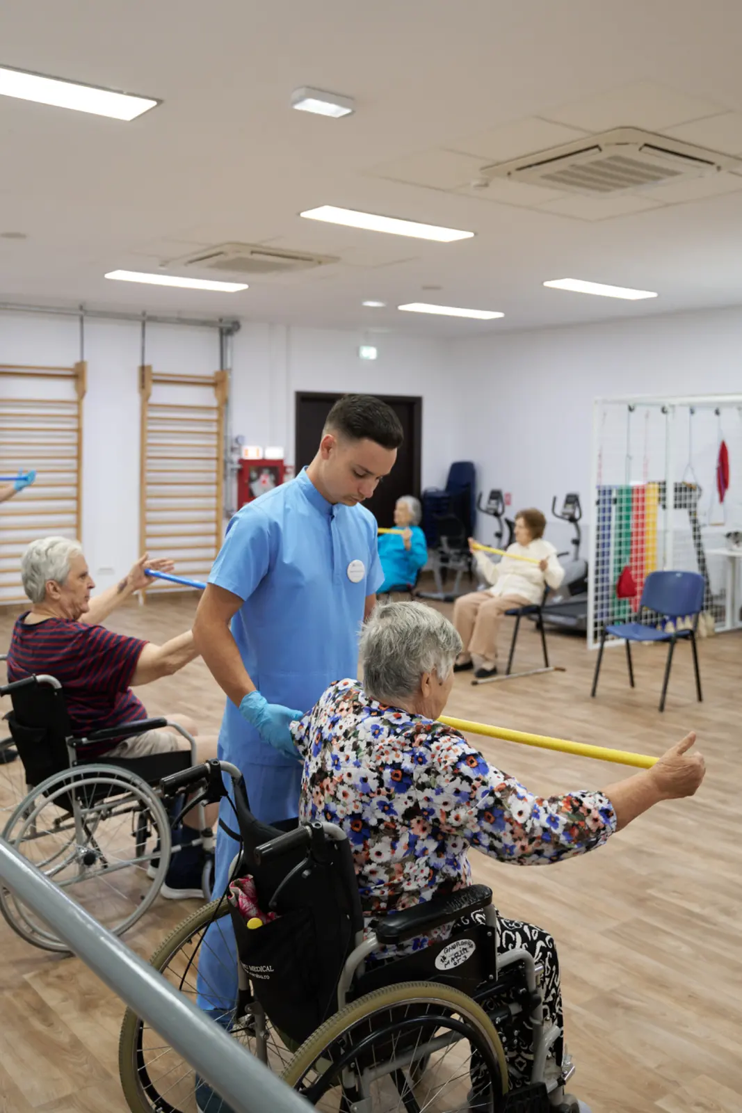 Kinetoterapeut în halat albastru ajutând o senioră în scaun rulant să efectueze exerciții cu baston galben, AsertivO Otopeni