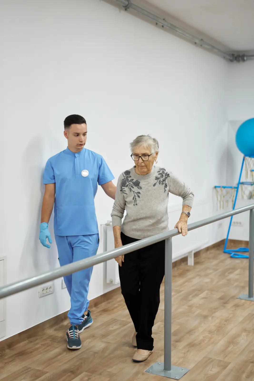 Kinetoterapeut în halat albastru sprijinind o senioră cu ochelari, AsertivO Otopeni
