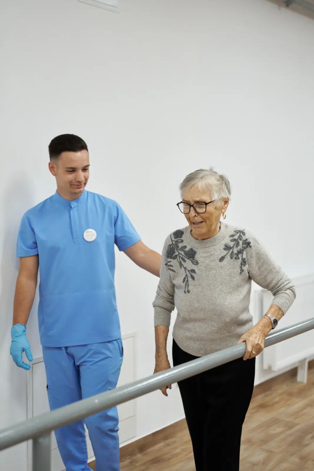 Kinetoterapeut în halat albastru sprijinind o senioră cu ochelari, AsertivO Otopeni