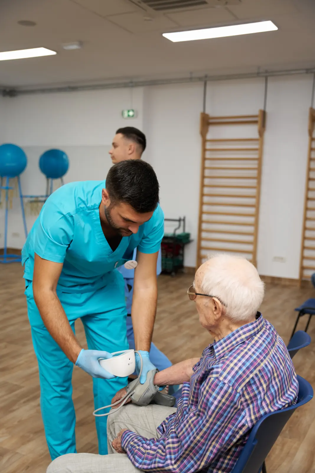 Kinetoterapeut în scrubs turcoaz măsoară tensiunea arterială unui senior cu păr alb, așezat pe scaun, AsertivO Otopeni
