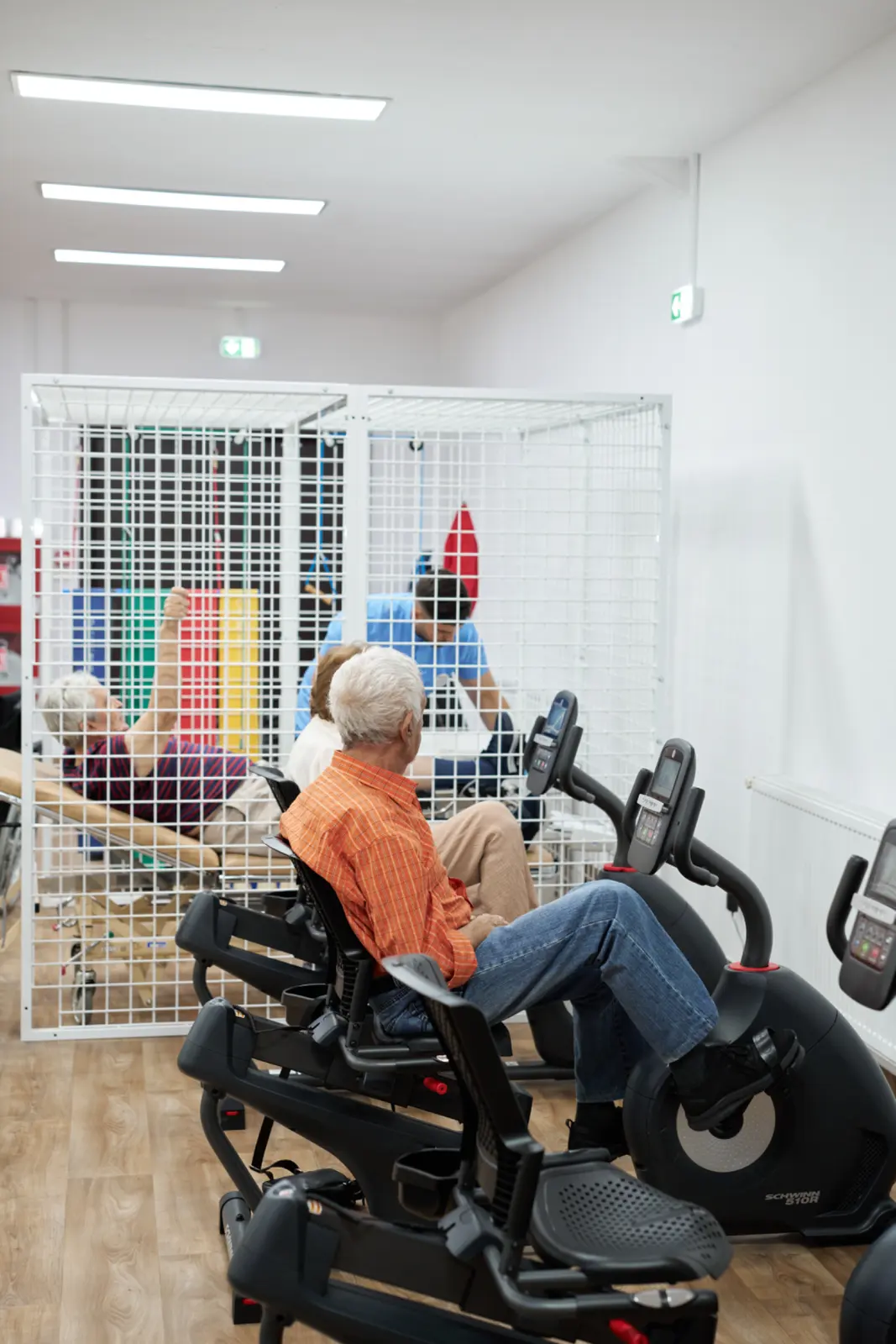 Senior în cămașă portocalie pe bicicletă ergometrică recumbentă într-o sală de kinetoterapie, AsertivO Otopeni