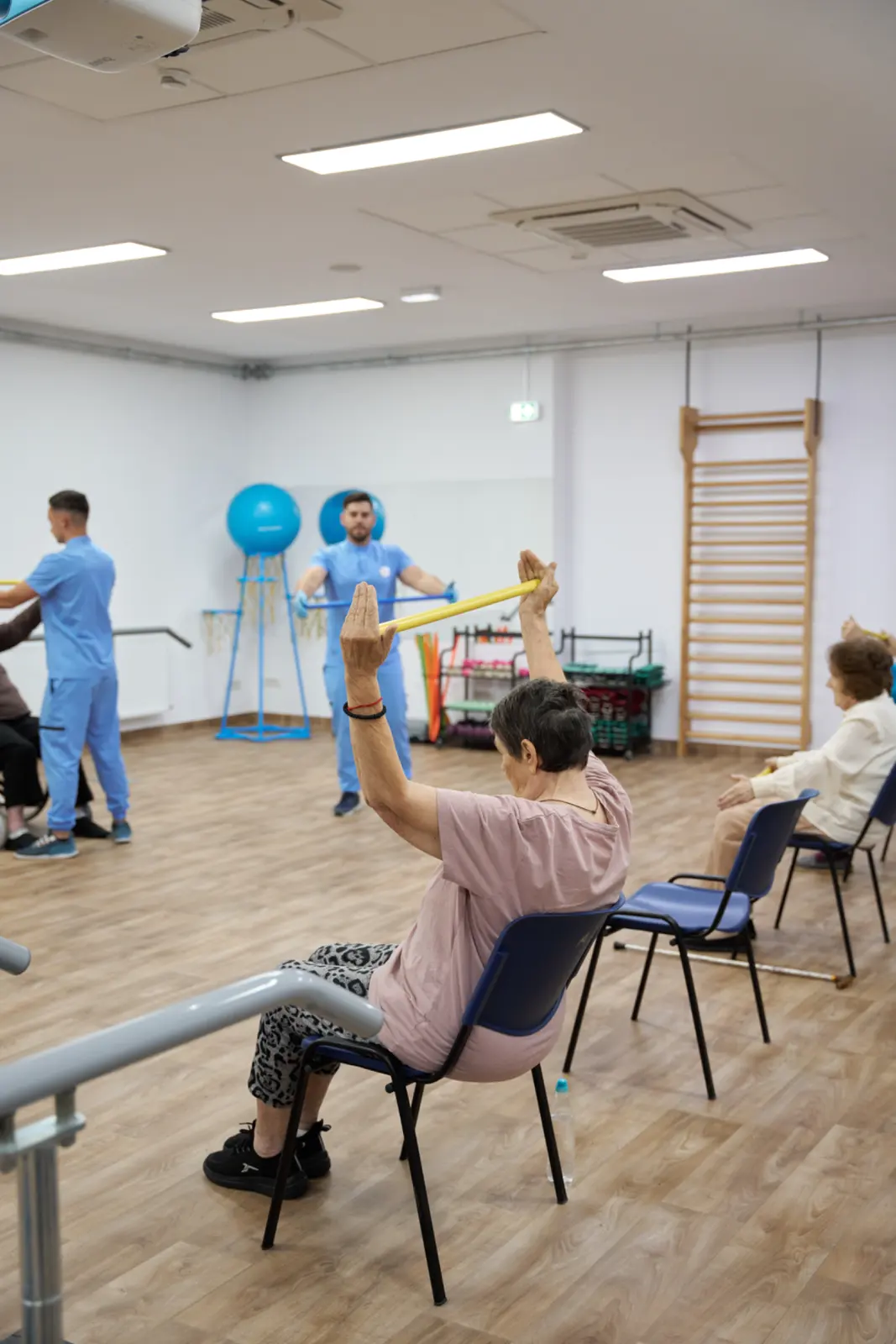 Seniori așezați pe scaune efectuând exerciții cu baston galben în sala de kinetoterapie, AsertivO Otopeni